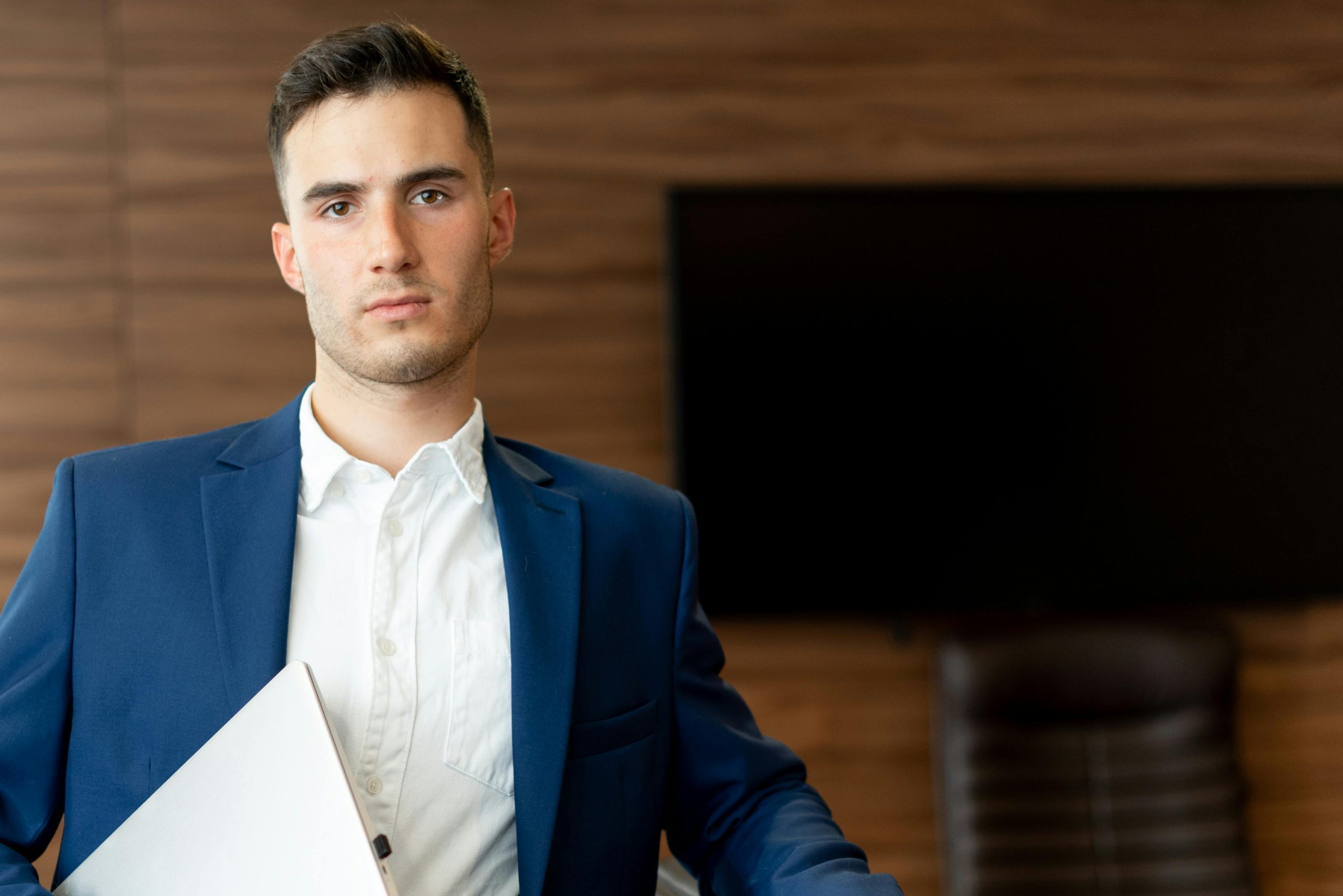 Confident young businessman in a blue suit, holding a laptop in an office setting.